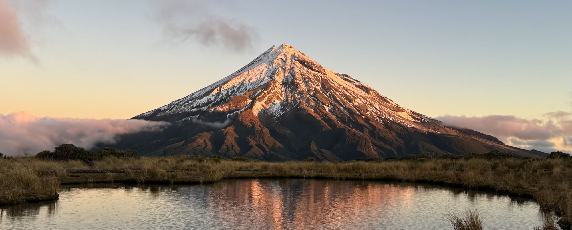 A snow-capped volcanic mountain lit by the evening sun, reflected in a still pool