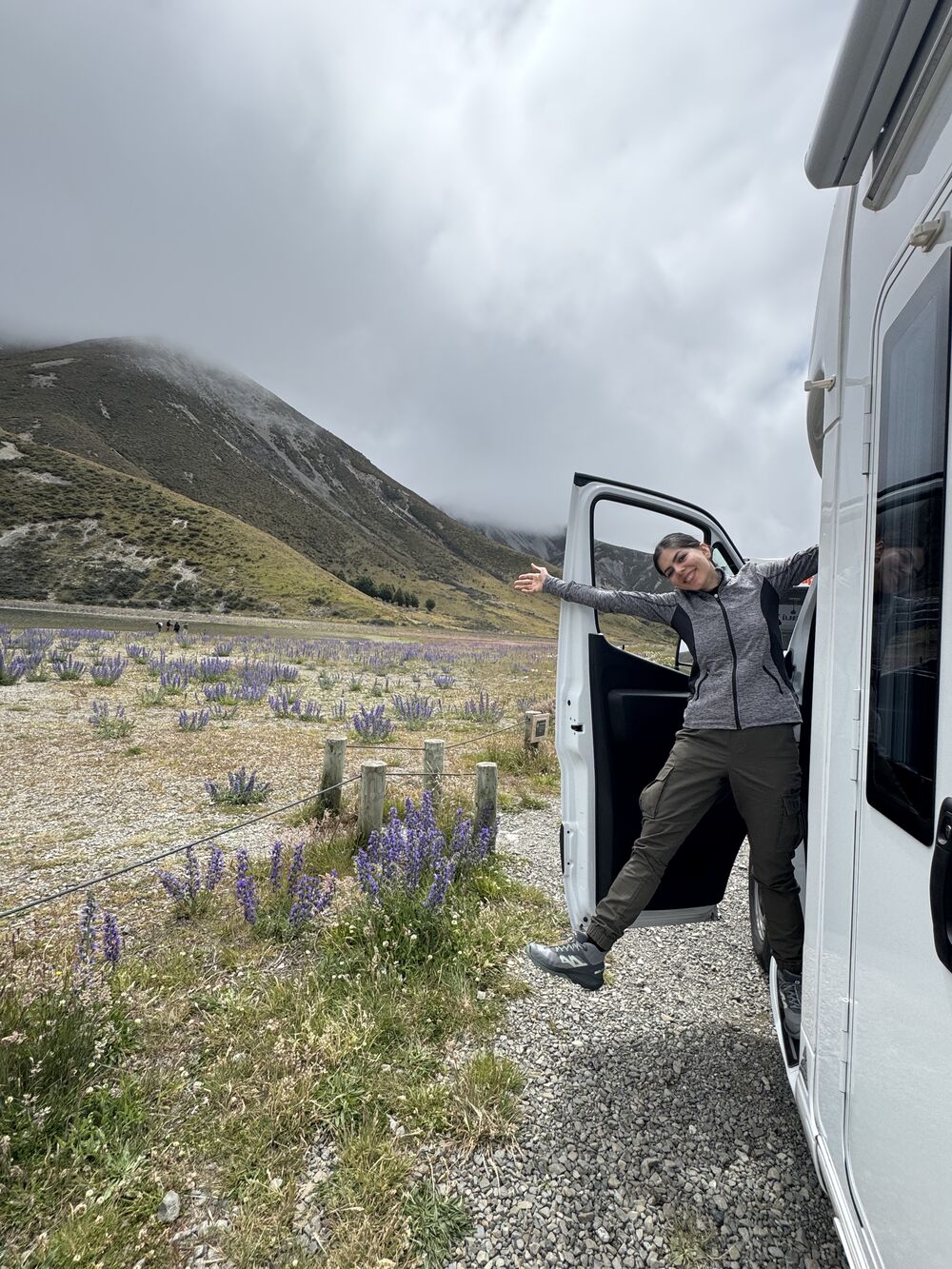 Mahnaz leaning out of a campervan door, smiling, beside a field of purple lupins and misty mountains