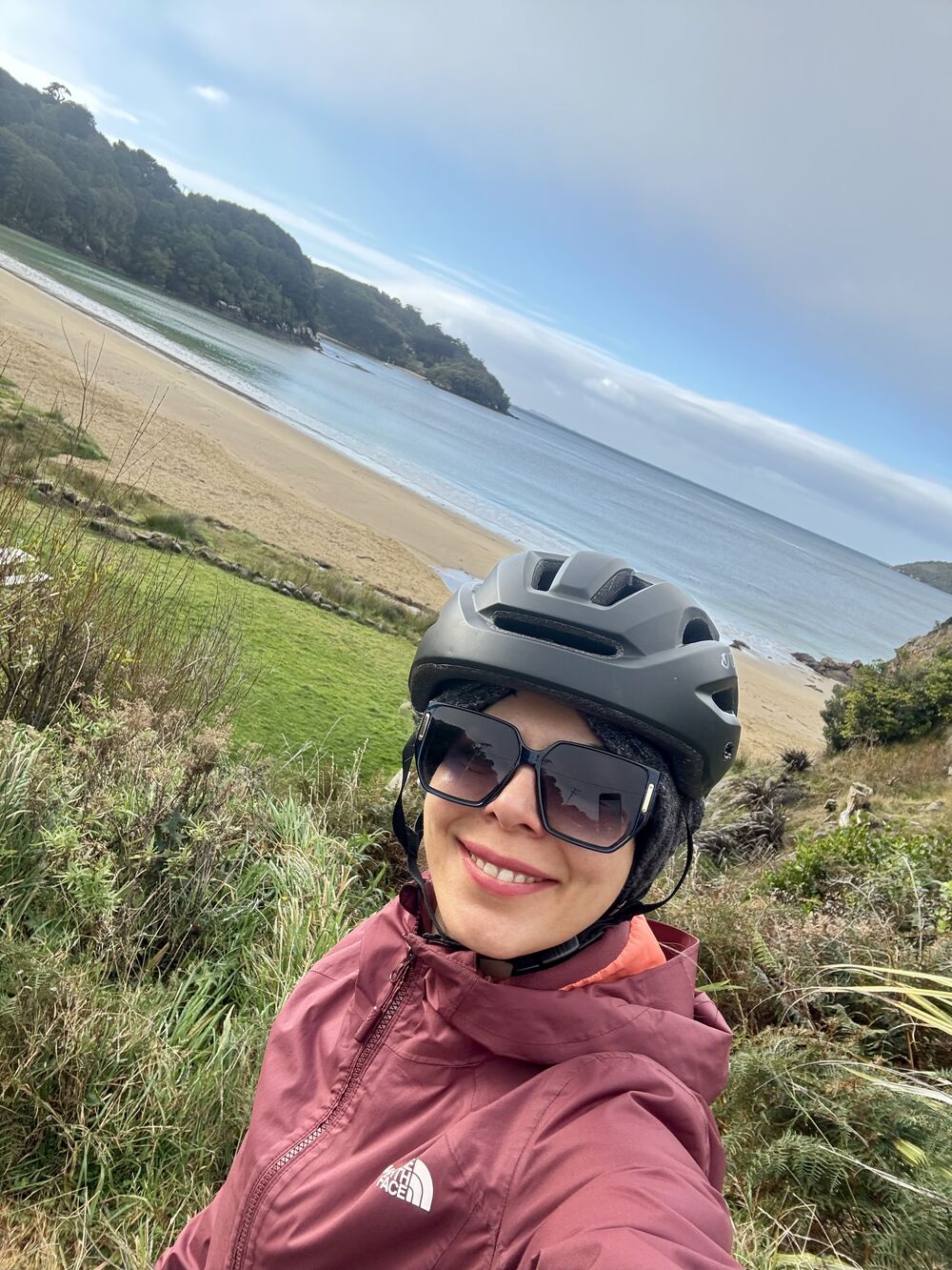 Mahnaz in a cycling helmet smiling, with a sandy bay behind