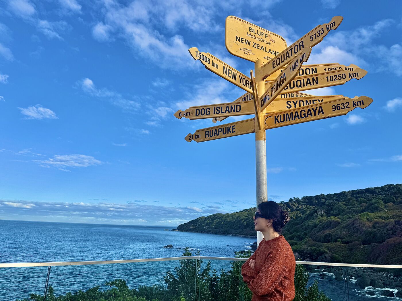 Mahnaz beside a yellow signpost pointing to distant cities, with the sea behind